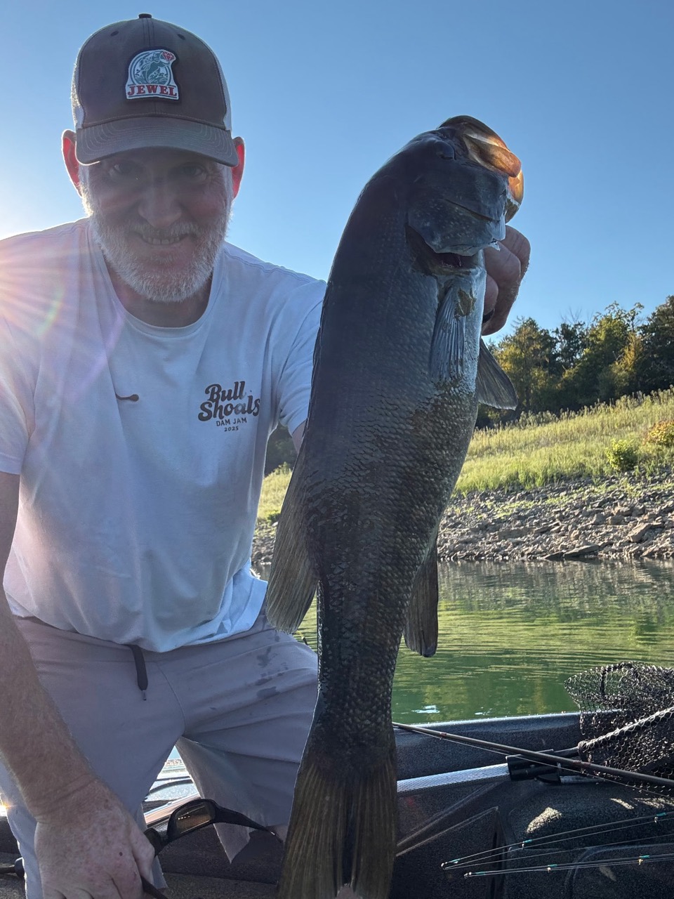 An angler holding a bass on a boat beside a wooded shoreline